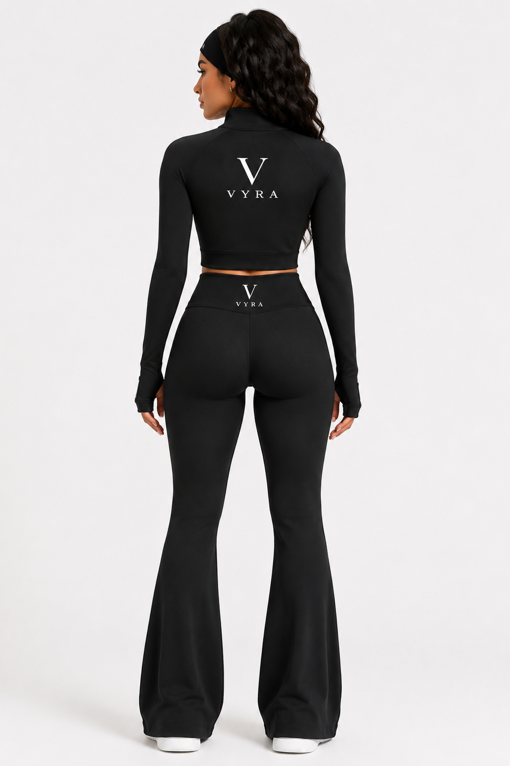 A woman shown from the back wearing a black long-sleeve crop top and matching high-waisted flared pants with a thin gold waistband. The brand name 'VYRA' is printed in white on the back of both garments.
