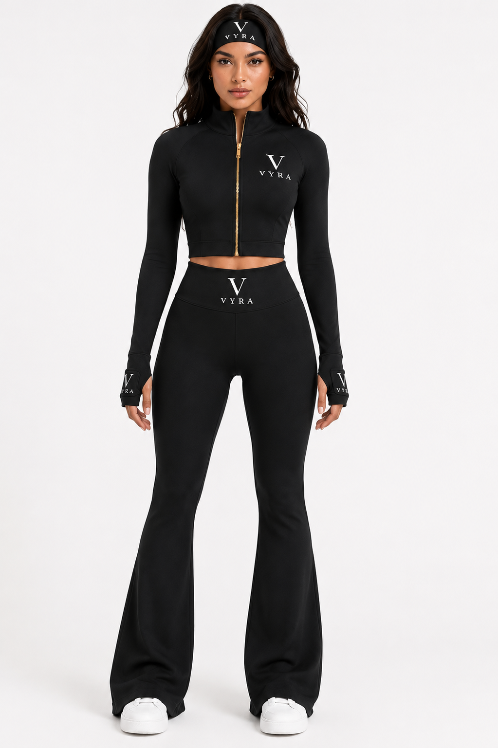 A model wearing a black two-piece athletic set consisting of a long-sleeve cropped zip-up top and matching high-waisted flared pants, both featuring a white Versace logo on the chest and waist.
She is also wearing a black Versace headband and white sneakers against a plain white studio background.
