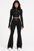 A model wearing a black two-piece athletic set consisting of a long-sleeve cropped zip-up top and matching high-waisted flared pants, both featuring a white Versace logo on the chest and waist.
She is also wearing a black Versace headband and white sneakers against a plain white studio background.