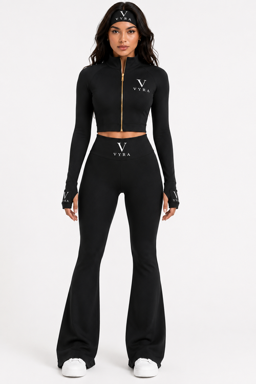 A model wearing a black two-piece athletic set consisting of a long-sleeve cropped zip-up top and matching high-waisted flared pants, both featuring a white Versace logo on the chest and waist.
She is also wearing a black Versace headband and white sneakers against a plain white studio background.