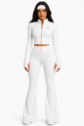 A woman modeling a white two-piece athletic set consisting of a cropped zip-up long-sleeve top and matching high-waisted flared pants, with pink 'Vibra' logos on the chest and cuffs, photographed against a solid white background.