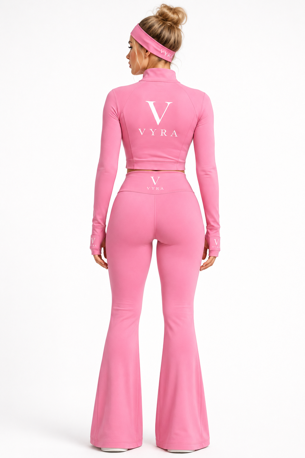 A woman shown from the back wearing a pink long-sleeve crop top and matching high-waisted flared leggings, with a pink headband. The outfit has a large white 'V' and 'VYRA' logo printed on the back of the top.