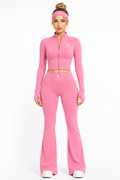 A model wearing a pink two-piece athletic set consisting of a long-sleeve crop top with a gold zipper and a matching high-waisted flare pant, along with a pink headband and white sneakers, 
on a white studio background