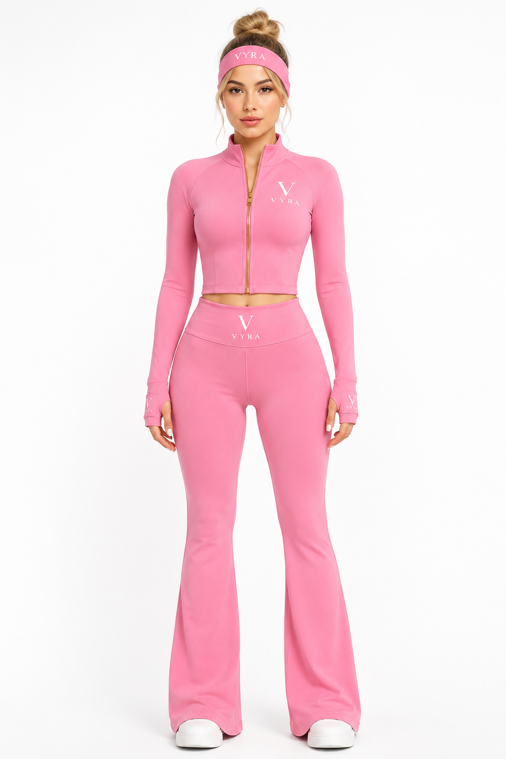 A model wearing a pink two-piece athletic set consisting of a long-sleeve crop top with a gold zipper and a matching high-waisted flare pant, along with a pink headband and white sneakers, 
on a white studio background